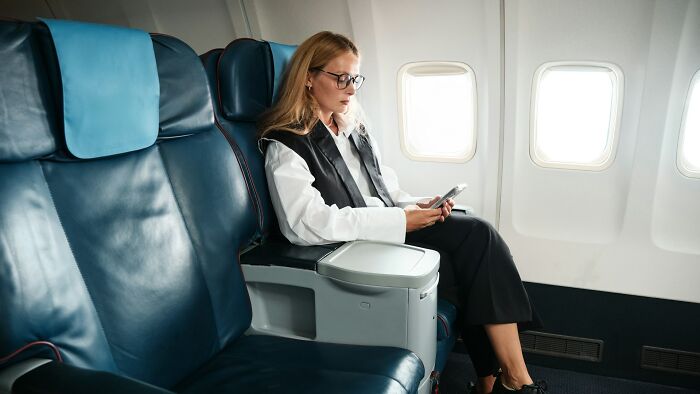 Woman sitting alone on airplane reading phone, illustrating weird and unhinged family traditions travel stories.