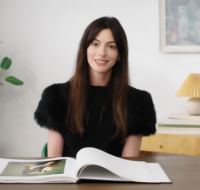 Anne Hathaway sitting at a table with an open book, wearing a black dress with puffed sleeves, smiling indoors.