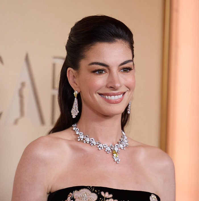 Anne Hathaway smiling at an event, wearing diamond earrings and a statement necklace, with hair styled back.