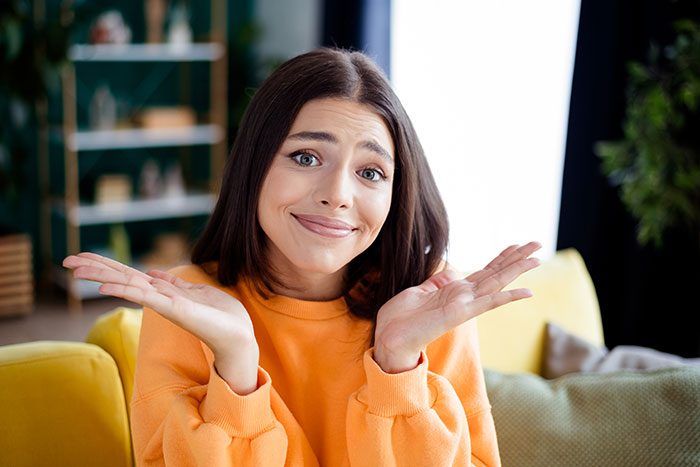Young woman in orange sweater shrugging indoors, expressing uncertainty about nephew's uncertain boxing match decision.