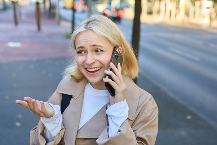 Woman talking on phone outdoors, smiling and gesturing, reflecting conflict over skipping nephew's boxing match for dogs.