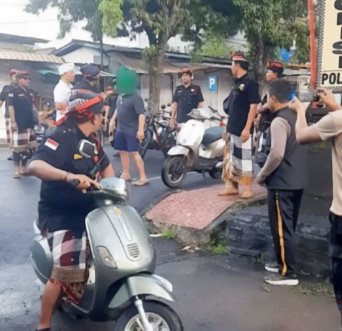 Indonesian police arrest American tourist during Bali shutdown as people and scooters gather on a wet street.