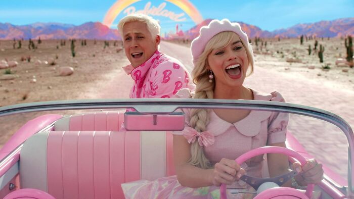 Two actors in bright pink outfits driving a vintage pink car in a desert setting with mountains in the background.