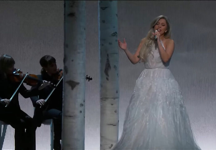 Female singer in a white gown performing alongside violinists in a staged setting for true stories that sound bizarre.