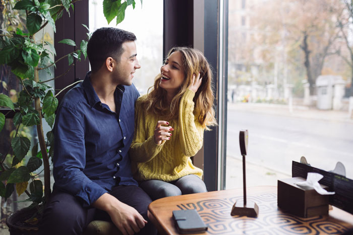 Couple sitting by a window in a caf&eacute;, boyfriend smiling and talking with woman, highlighting fatherhood and medical procedure concerns.