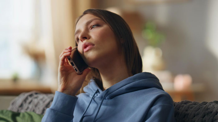 Young woman in a blue hoodie talking on the phone looking thoughtful in a cozy home setting, best friend fight mom topic