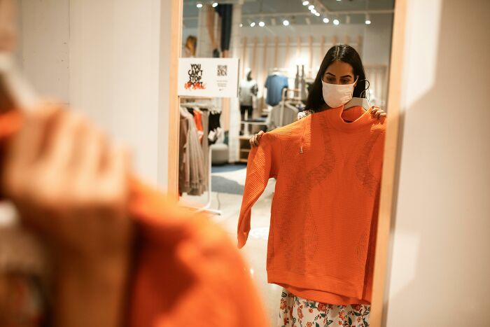 Woman wearing mask holding an orange sweater in front of a mirror inside a clothing store, illustrating epic PR fails concept.