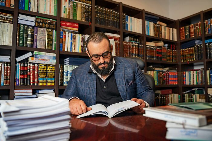 Man in glasses and blazer reading a book in a library filled with legal and PR fail related books on shelves.