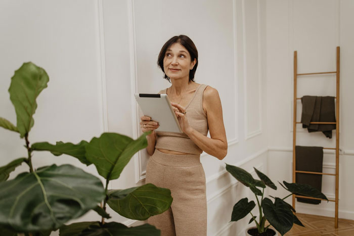 Woman playing dorm inspector for son's boyfriend, checking out the diggs with a tablet in a stylish, plant-filled room.