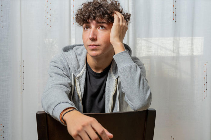 Young man sitting indoors looking thoughtful as mom plays dorm inspector checking boyfriend&rsquo;s digs before moving in