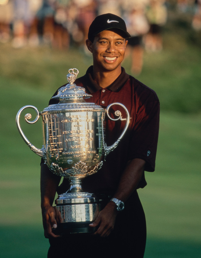Tiger Woods segurando um grande troféu de golfe, sorrindo e vestindo um boné preto e uma camisa marrom em um campo de golfe. Tiger Woods segurando um grande troféu de golfe, sorrindo e vestindo um boné preto e uma camisa marrom em um campo de golfe.