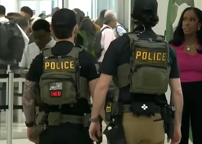 Two police officers in tactical gear walking through a busy airport crowd during an ICE encounter.