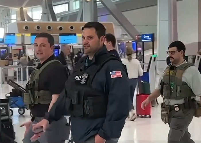 Three law enforcement officers in tactical gear at an airport capturing attention with a Brooklyn man’s reaction to ICE.