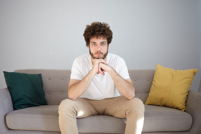 A young man with curly hair and a beard sits on a couch, looking forward, deep in thought about his proposal.