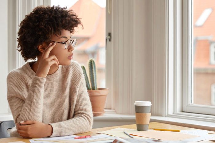 Young woman with glasses sitting by window, looking thoughtful about brother inherited land money and documents on table.