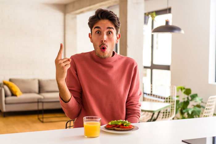 Young man in a pink sweater seated at a table with breakfast, expressing surprise and pointing upwards in a bright room.