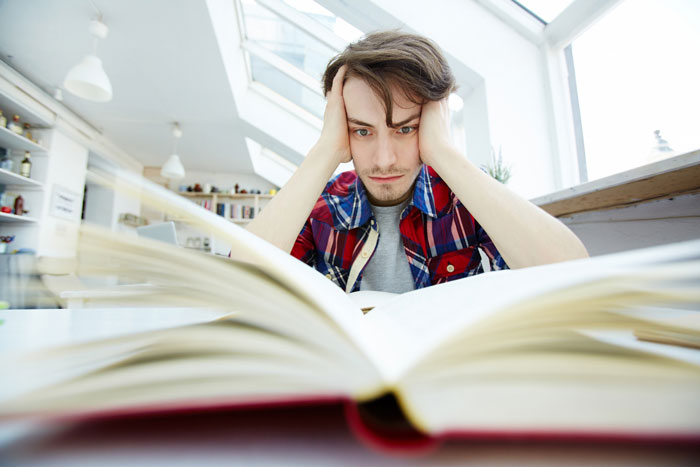 Frustrated young man reading a large book indoors, representing jobless struggle and family competition with PhD cousin.