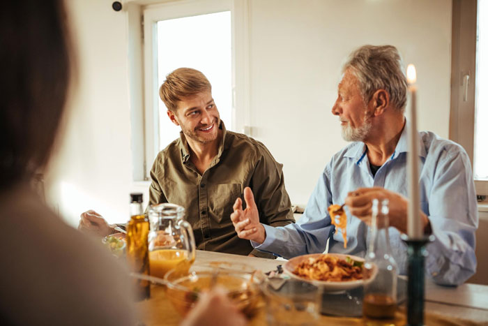 Young man and elderly man discussing with concerned expressions during a family meal about jobless competition and life savings loss.
