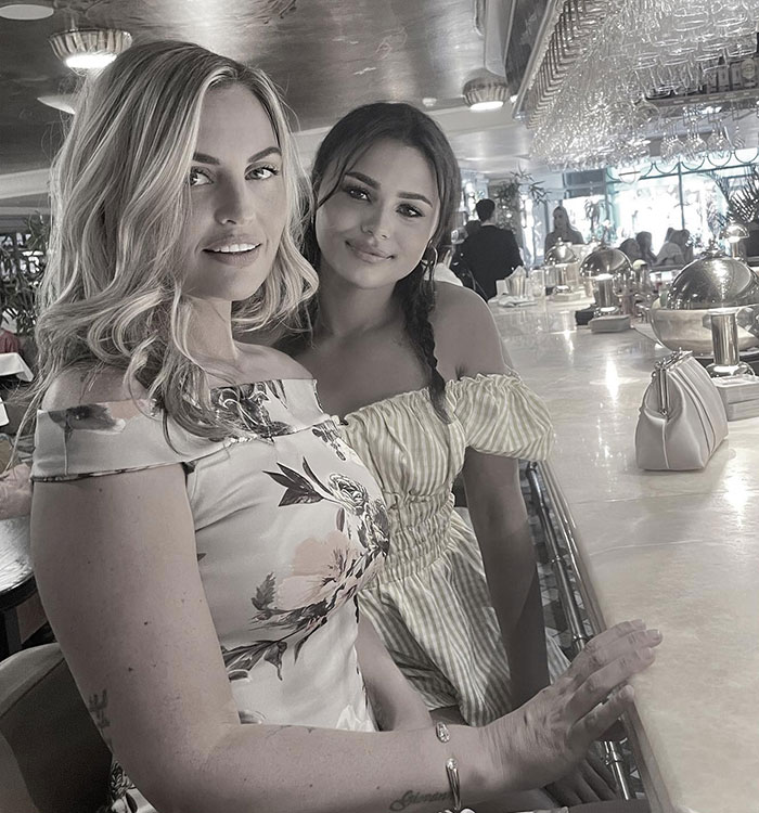 Two women sitting at a bar, one with blonde hair and floral dress, the other with dark hair in a striped off-shoulder dress.