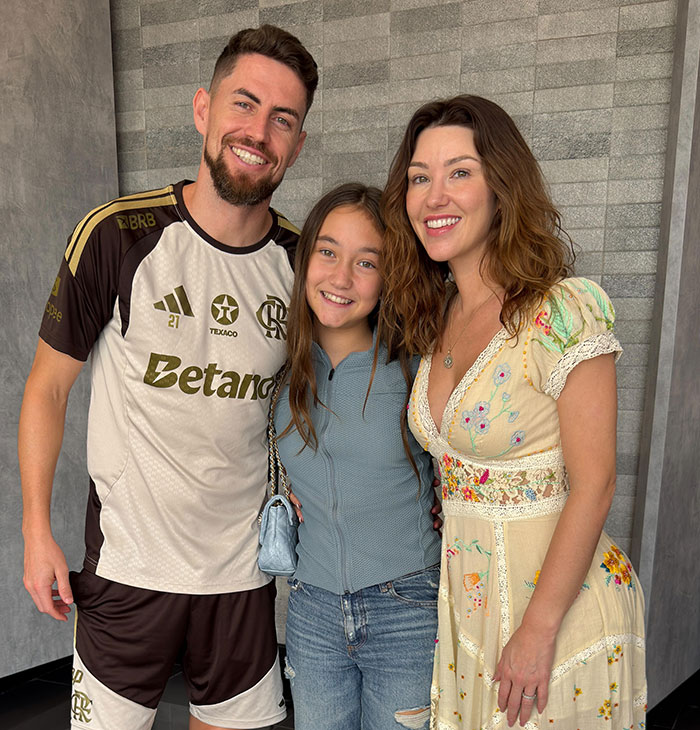 Security guard at the center of Chappell Roan debacle smiling with two women in casual attire against a gray wall background.