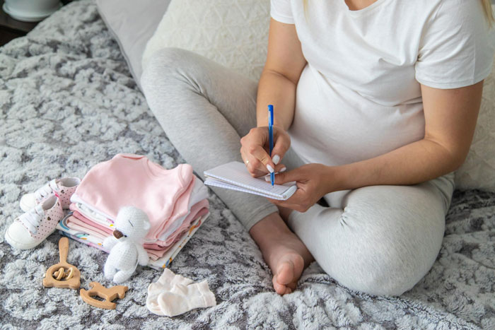 Pregnant woman sitting on bed writing in notebook next to baby clothes and toys, changing baby name secretly concept.