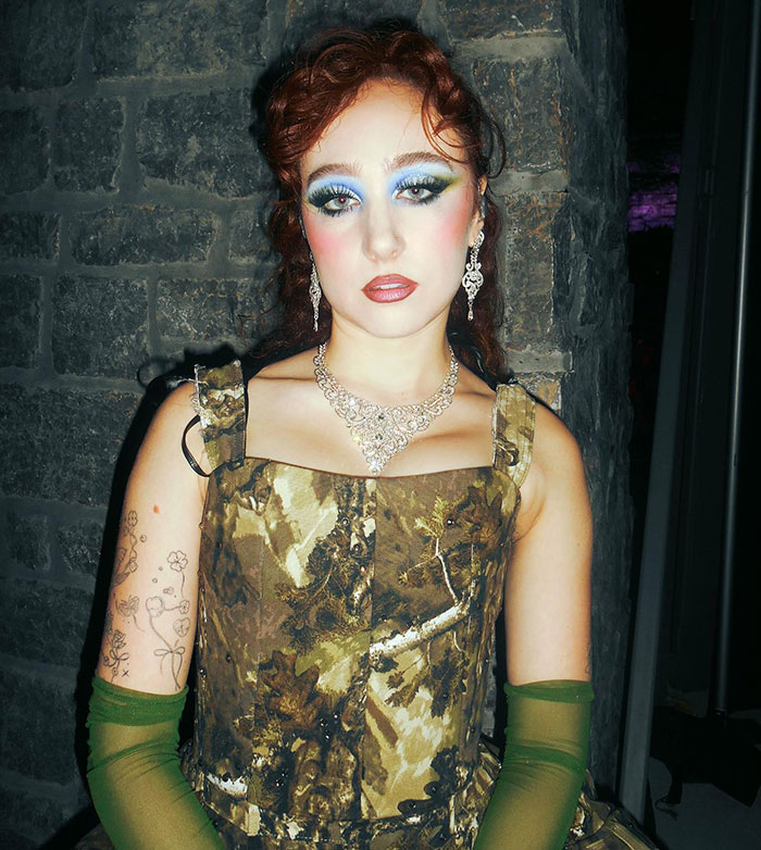 Chappell Roan wearing green gloves and earrings, posing against a stone wall with dramatic makeup and jewelry.