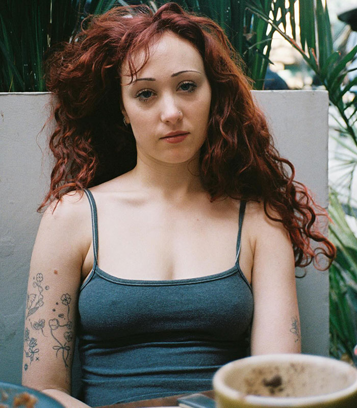 Chappell Roan with curly red hair and tattoos, wearing a gray tank top, sitting indoors with plants in the background Chappell Roan with curly red hair and tattoos, wearing a gray tank top, sitting indoors with plants in the background