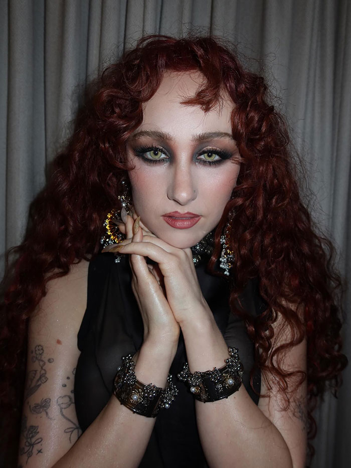 Chappell Roan with curly red hair and dramatic makeup posing with hands near face against a curtain backdrop.