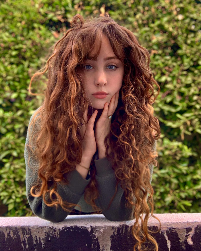 Chappell Roan with long curly hair, resting face on hands, posing outdoors against green leafy background.
