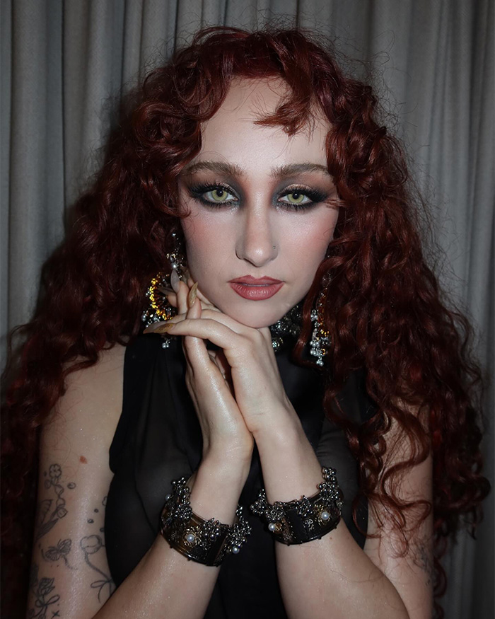 Chappell Roan with red curly hair and dramatic makeup, posing with hands near face amid backlash news. Chappell Roan with red curly hair and dramatic makeup, posing with hands near face amid backlash news.