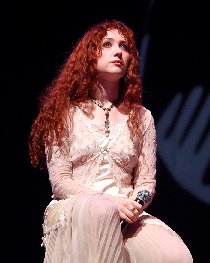 Chappell Roan seated on stage holding microphone, wearing a light pink outfit with long red curly hair. Chappell Roan seated on stage holding microphone, wearing a light pink outfit with long red curly hair.