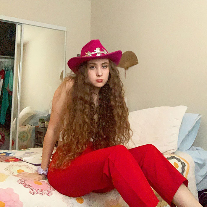 Chappell Roan sitting on a bed wearing a pink cowboy hat and red pants with long curly hair in a casual room setting