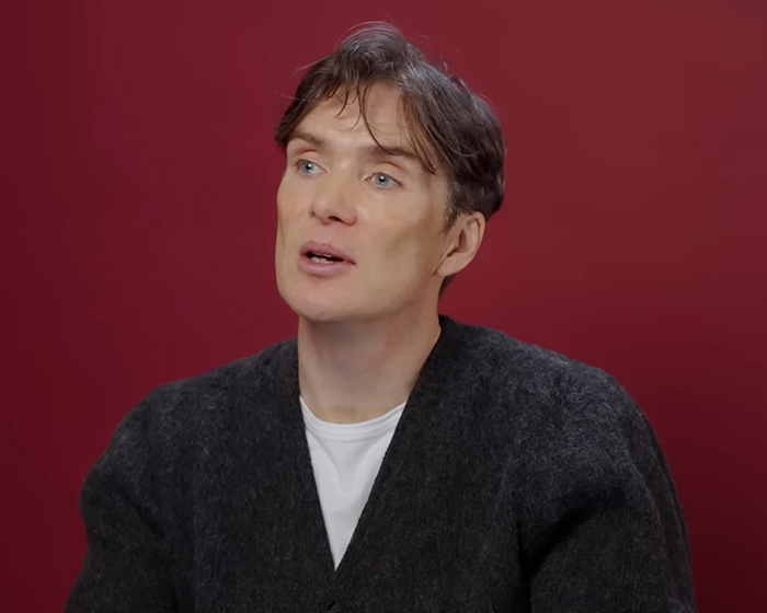 Cillian Murphy speaking against a red background, wearing a dark sweater, sparking debate about his aging appearance.