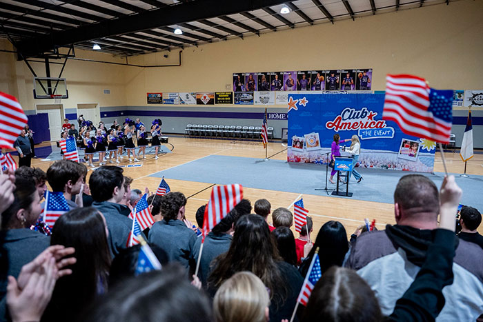 Crowd waving American flags at an indoor event with a stage backdrop featuring Club America branding.