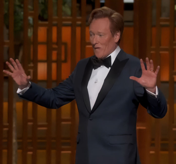 Comediante no palco do Oscar 2026, envolvendo o público com linguagem corporal expressiva e gestos abertos. Comediante no palco do Oscar 2026, envolvendo o público com linguagem corporal expressiva e gestos abertos.