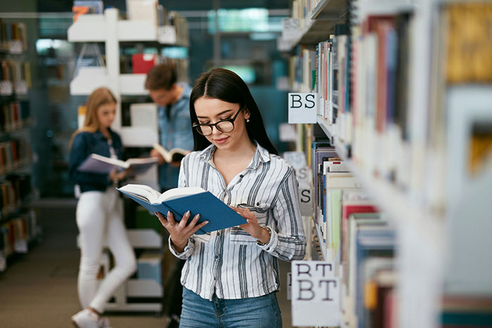 Young woman reading a book in a library with shelves in the background, related to businesses and corruption topics.