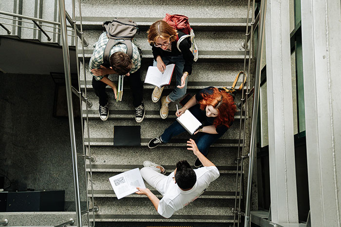 Four young people studying and discussing business corruption on a stairwell with books and laptops around them