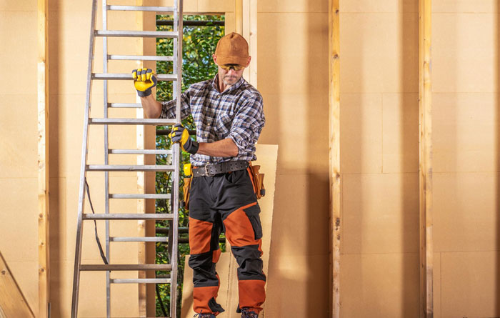 Man in workwear carrying a ladder inside a wooden frame structure, illustrating lazy guy stealing coworker&rsquo;s ladder concept.