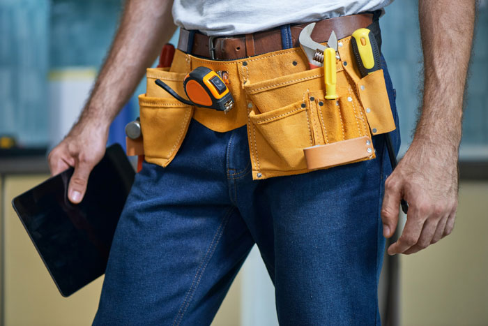 Man wearing tool belt with various tools including wrench, screwdriver, and tape measure while holding a digital tablet.