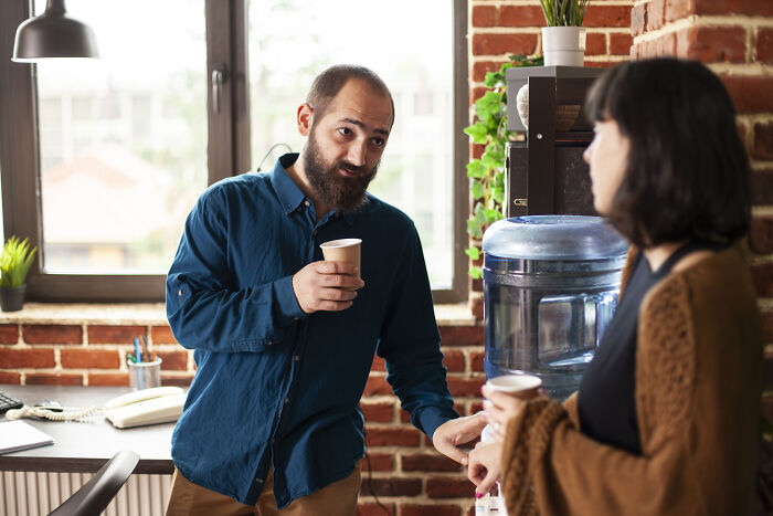 Man in blue shirt mansplaining to a woman by the water cooler in a casual office with exposed brick walls.