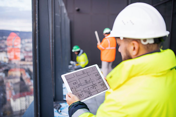 Construction worker in safety gear reviewing building plans on tablet while coworkers work in background on site.