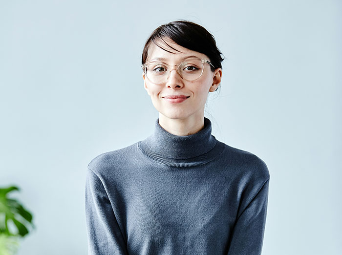 Young woman wearing glasses and a gray turtleneck smiling confidently in an office setting, related to coworker revenge stories.