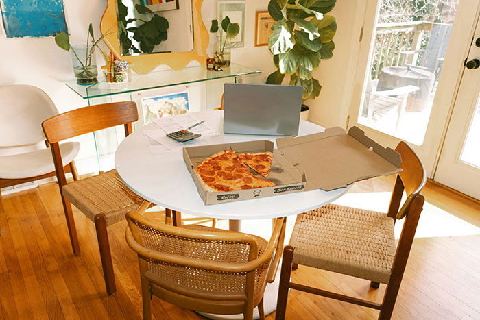 A home office setup with a laptop, papers, and a half-eaten pizza on a round table with wooden chairs.
