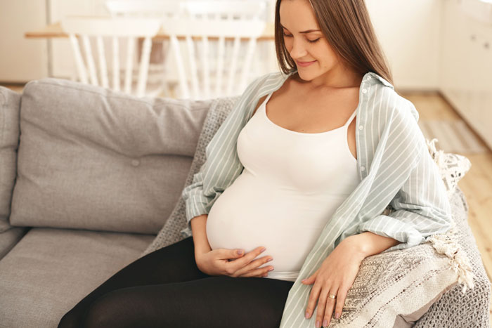 Pregnant daughter sitting on couch, gently holding belly, symbolizing concerns of home turned landfill and CPS involvement.