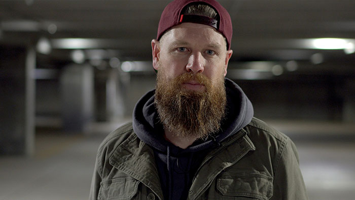 Bearded man wearing a red cap and green jacket in a dimly lit parking garage, representing caught cheating stories.