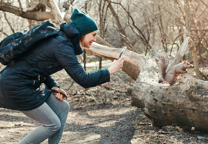 Woman wearing teal beanie and black jacket capturing a close photo of a squirrel in the woods showing displays of intelligence.