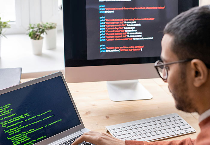 Person wearing glasses coding on laptop and desktop screens showing programming scripts, displaying intelligence and focus.