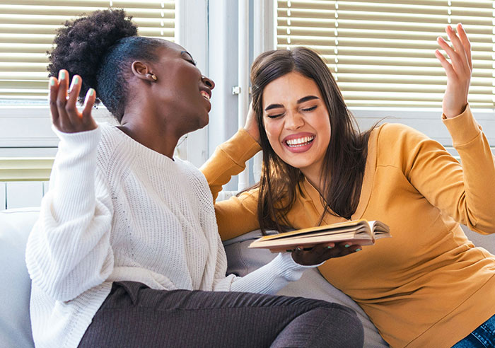 Two women laughing and enjoying a conversation while sharing a book, showcasing displays of intelligence.
