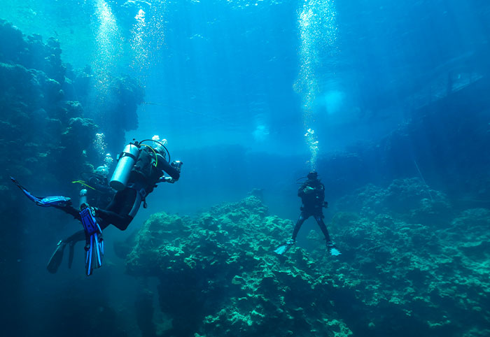 Two scuba divers exploring deep ocean waters above rocky coral formations with bubbles rising toward the surface.
