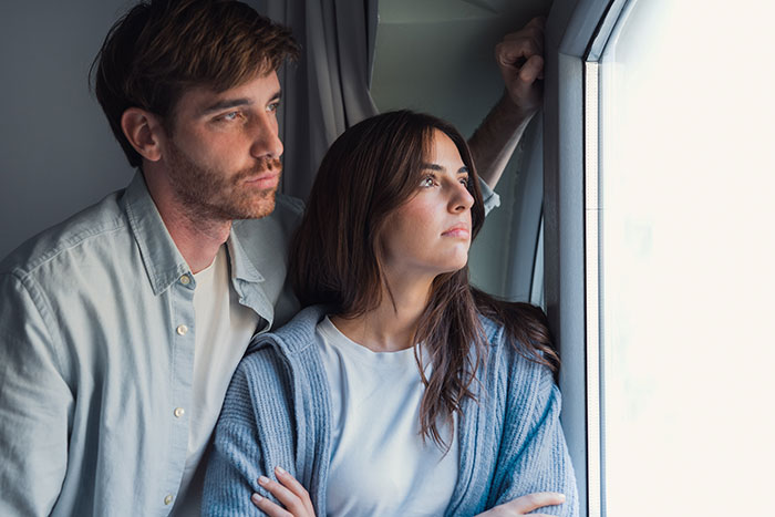 Man and woman looking out window, appearing worried and distressed over vandalized daughter's grave and vile letters.